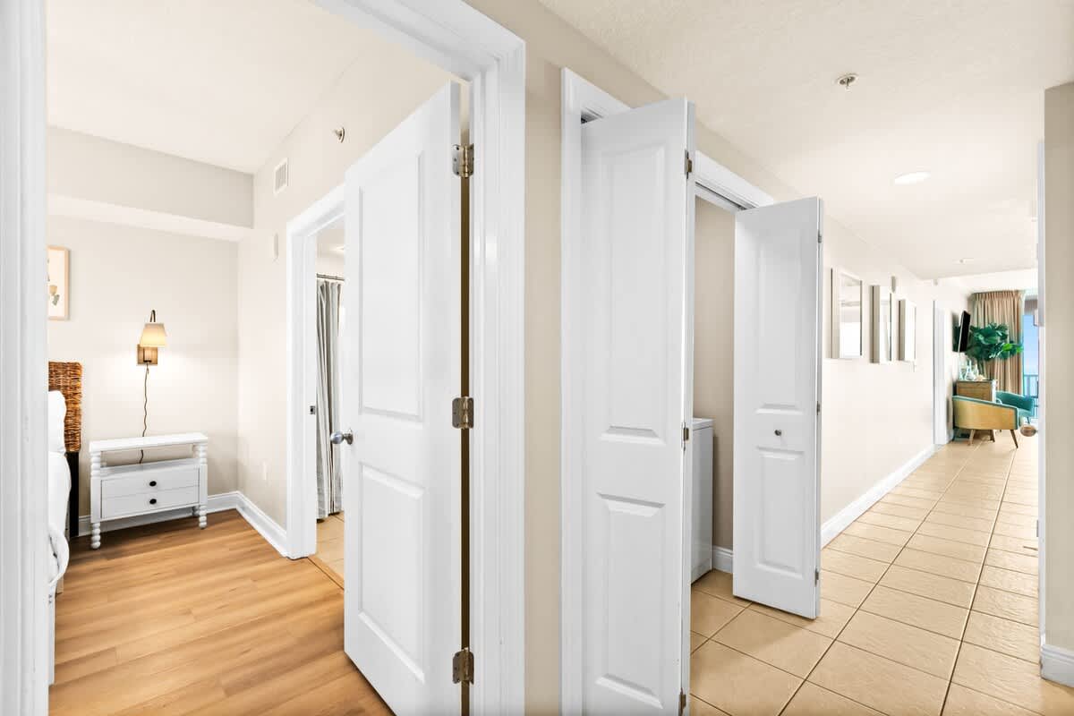 A hallway with light tile flooring and multiple white doors, providing a glimpse into different rooms, including a bedroom with wood flooring and a laundry area.