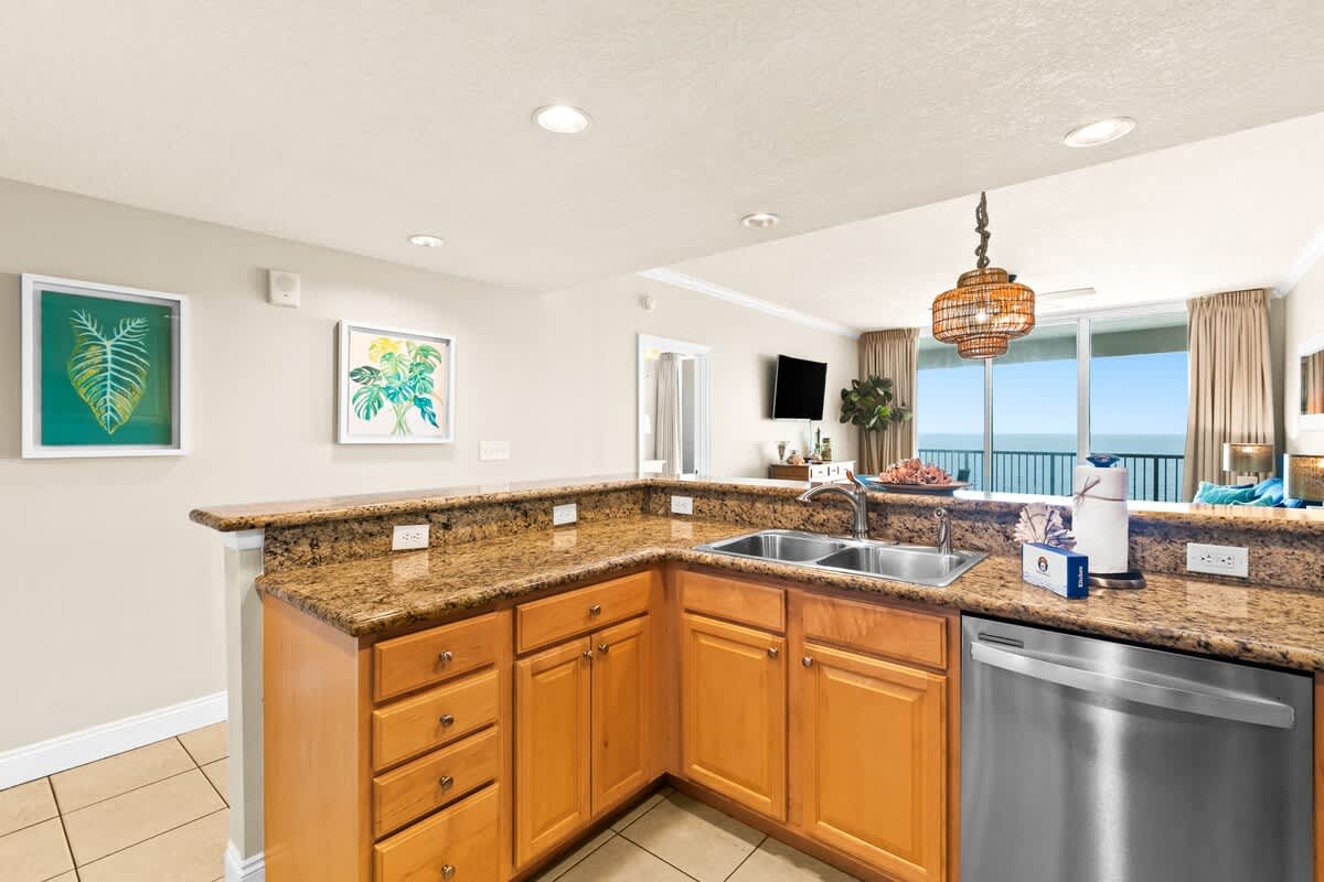 A bright kitchen with light wood cabinetry, speckled granite countertops, and stainless steel appliances, featuring a double sink and a breakfast bar overlooking the living area and ocean views.