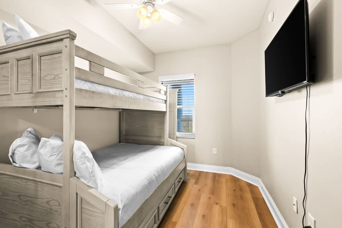 A cozy bunk room features a light grey wooden bunk bed with a full-size bottom bunk and a twin top bunk. A wall-mounted TV and a small window with blinds are also visible.