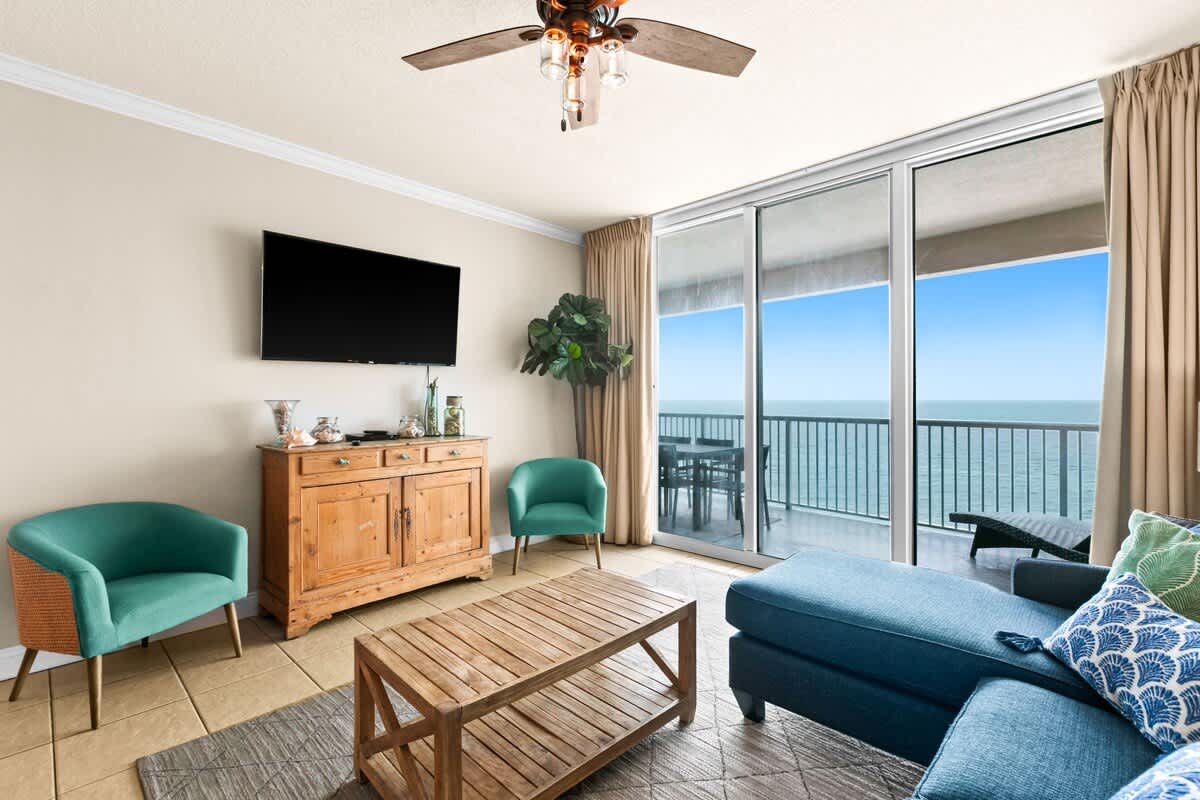 Inviting living room with a wall-mounted flat-screen TV above a rustic wooden console. A blue sectional sofa and two teal accent chairs surround a wooden coffee table, all facing sliding glass doors to an ocean-view balcony.