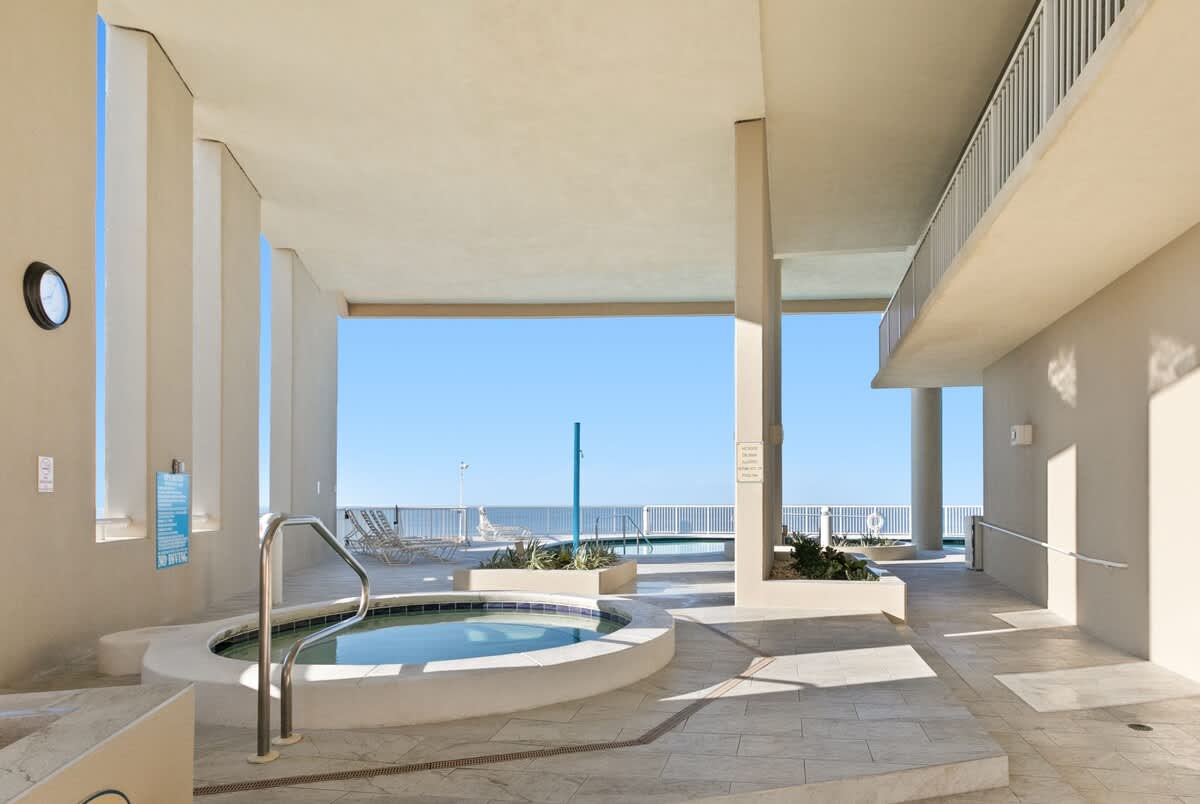 A circular outdoor hot tub with stainless steel handrails, surrounded by light-colored tile, located under a building overhang with glimpses of the ocean in the background.