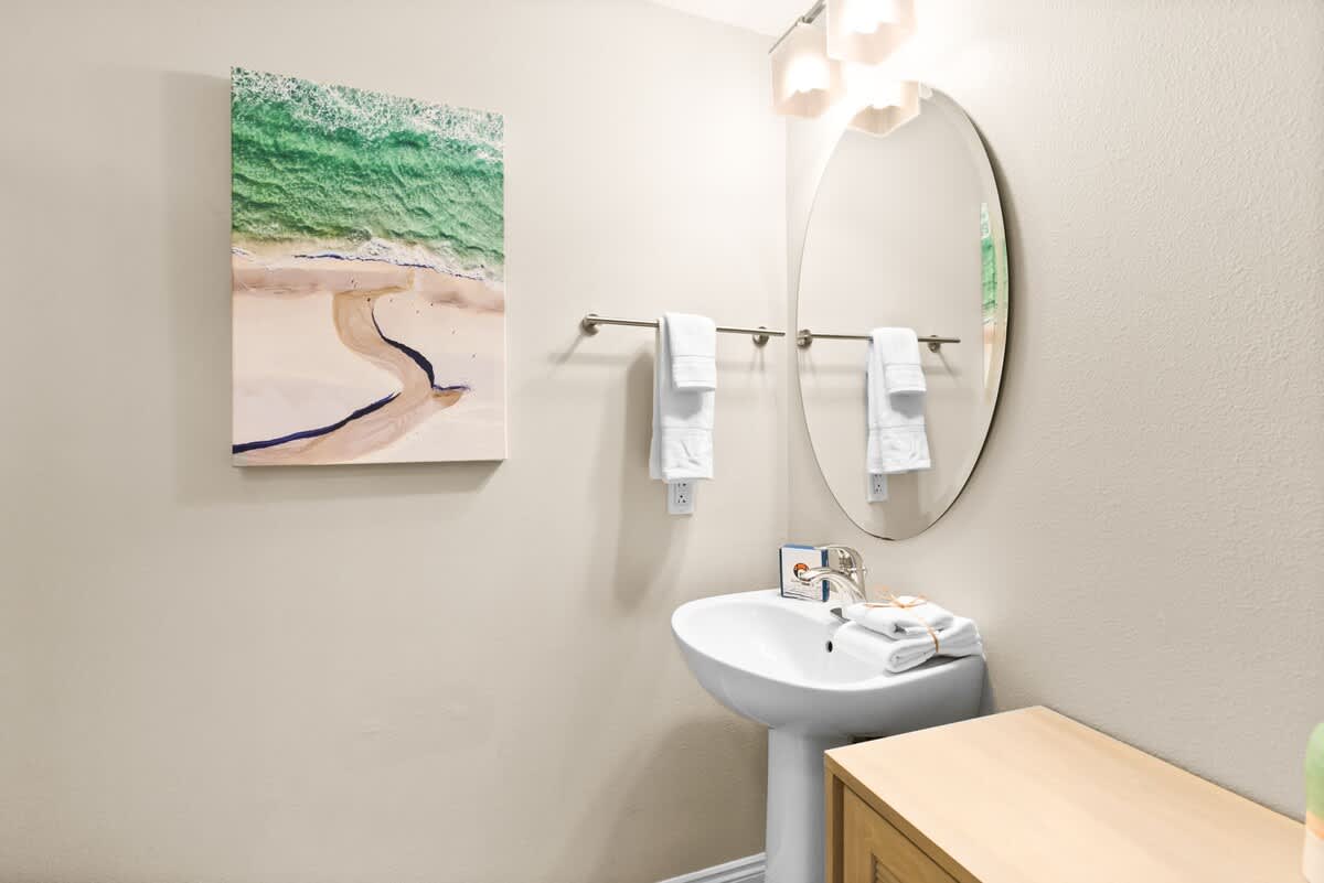 The bathroom features a white pedestal sink with a large oval mirror and modern light fixture above it. A colorful aerial beach painting adds a touch of coastal decor.
