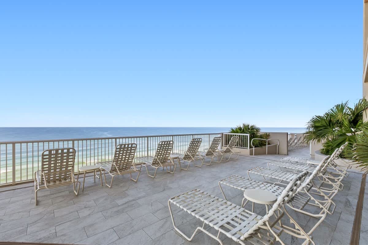 A spacious outdoor deck furnished with numerous white lounge chairs, offering an unobstructed, panoramic view of the blue ocean and sky, with lush palm foliage on the side.