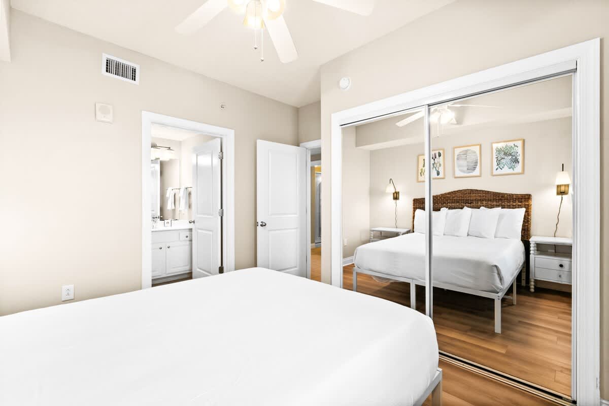 A bright bedroom features a white bed, a ceiling fan, and a large mirrored closet reflecting the room. An open doorway leads to an en-suite bathroom with a white vanity.