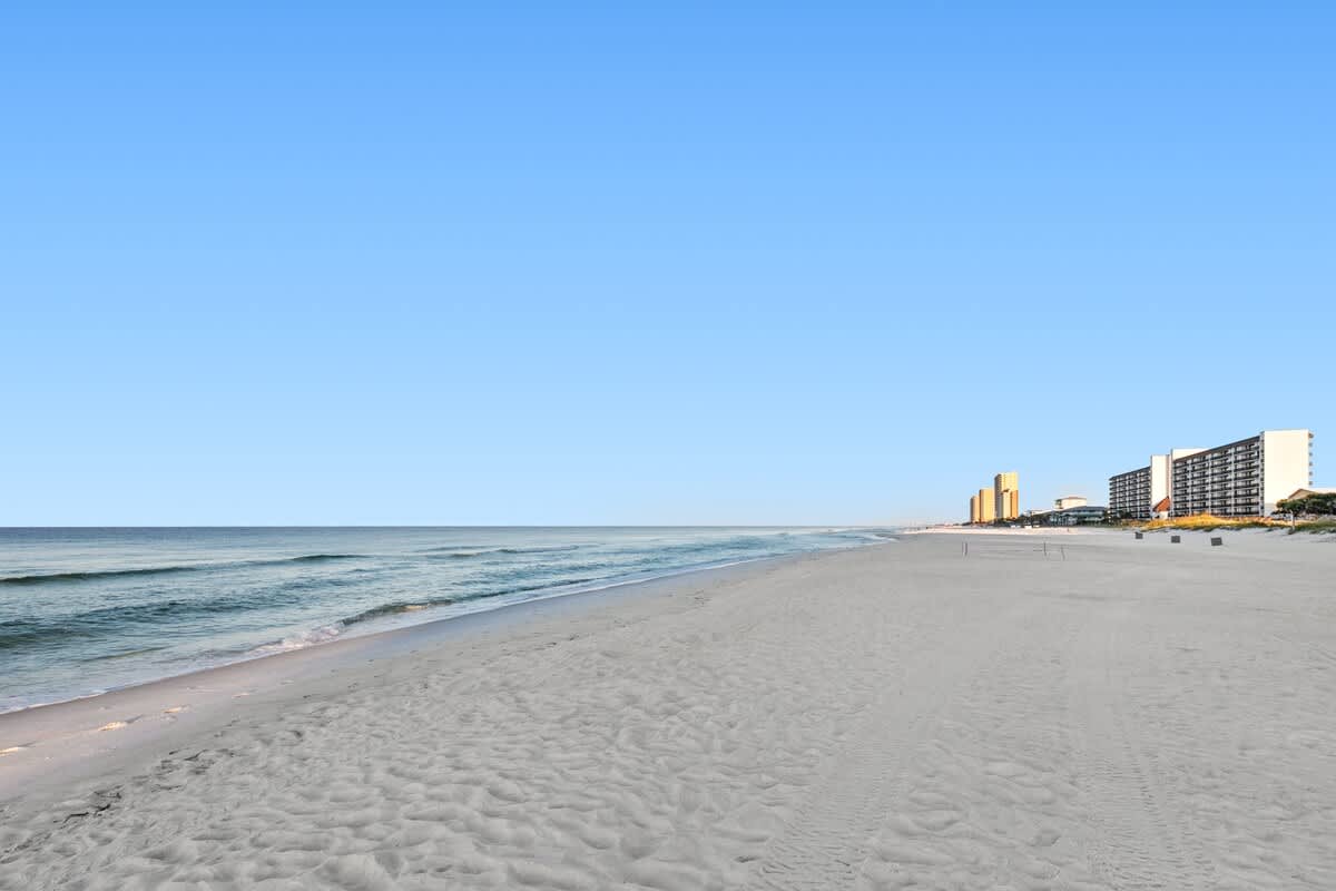 A vast expanse of white sand beach stretches along the coastline, meeting the turquoise ocean under a clear blue sky. Distant high-rise buildings are visible on the right horizon.