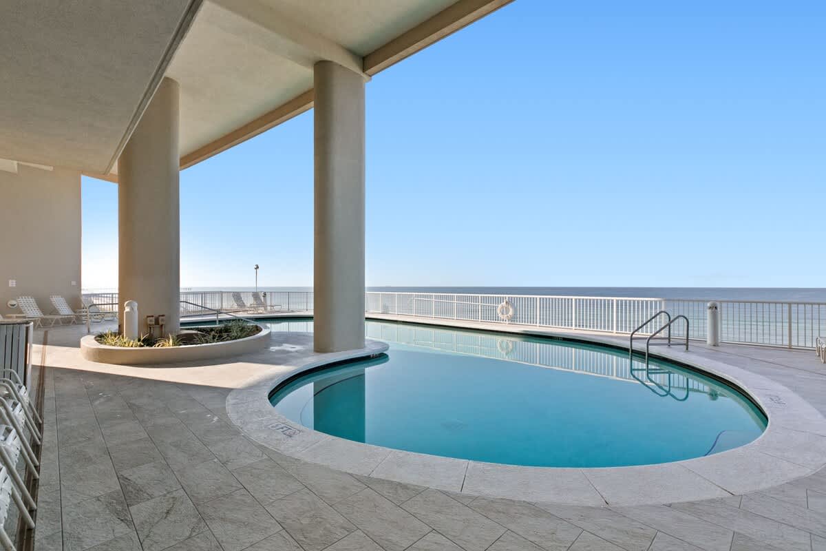 A communal pool area with a kidney-shaped pool, surrounded by light-colored tile, offering expansive views of the clear blue ocean and sky, with lounge chairs visible.