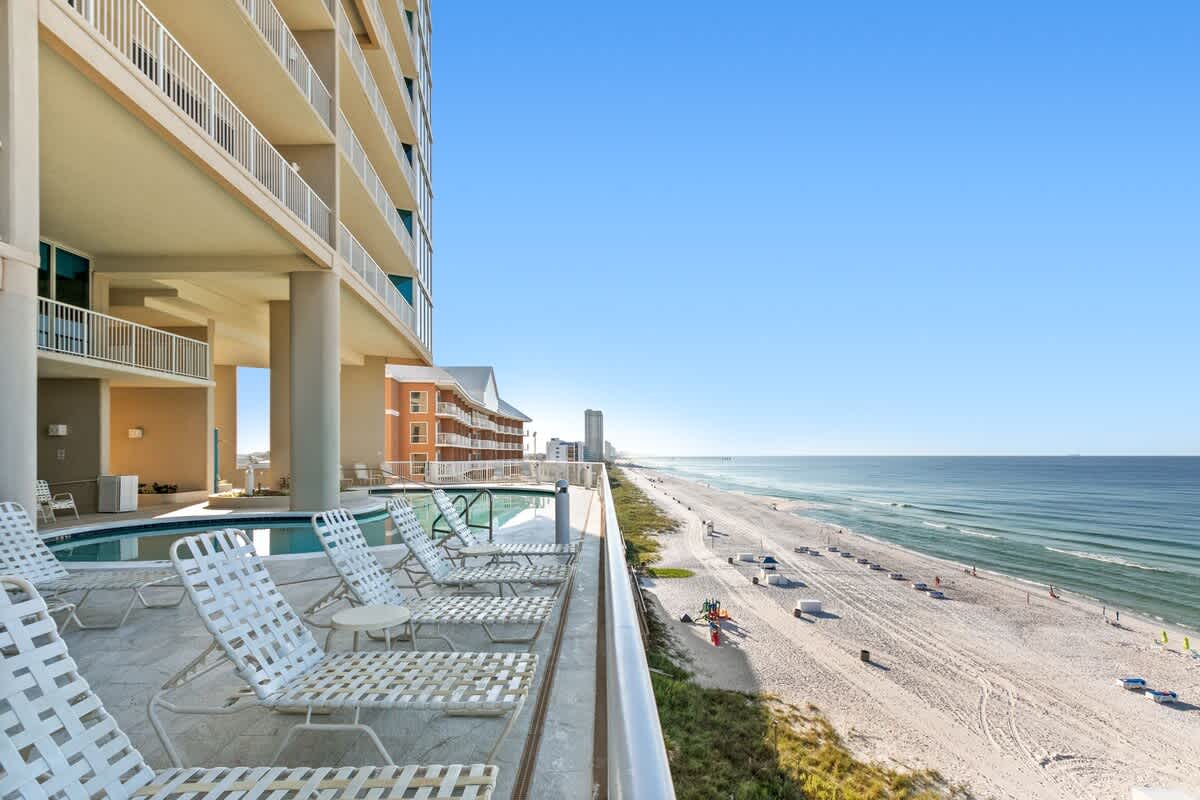 An elevated outdoor deck area with white lounge chairs and a communal pool, offering panoramic views of a wide sandy beach and the clear blue ocean stretching to the horizon.