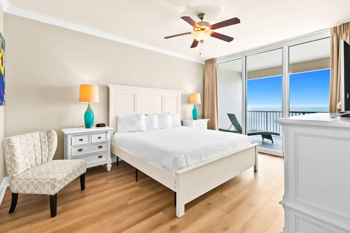 A bright bedroom featuring a white headboard, two nightstands with lamps, and a patterned accent chair, with large sliding glass doors offering expansive ocean views from the balcony.