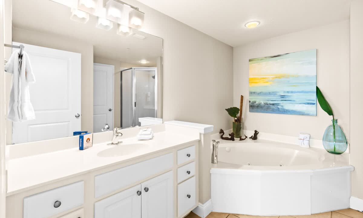 A spacious bathroom featuring a white jetted corner tub, a glass-enclosed shower, and a vanity with a large mirror, all set on light tile flooring.
