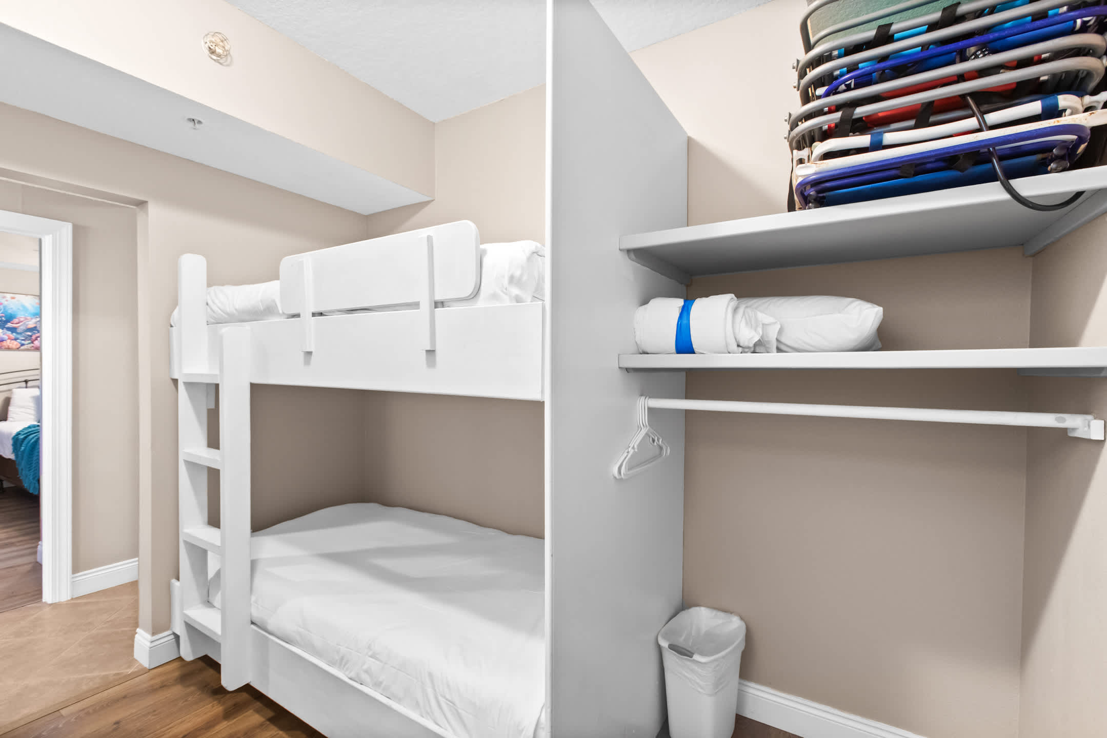 A white wooden bunk bed with white bedding is visible on the left, while a closet on the right features shelves stacked with beach chairs and a rolled-up mattress or pillow. The room has light beige walls and wood-look flooring.