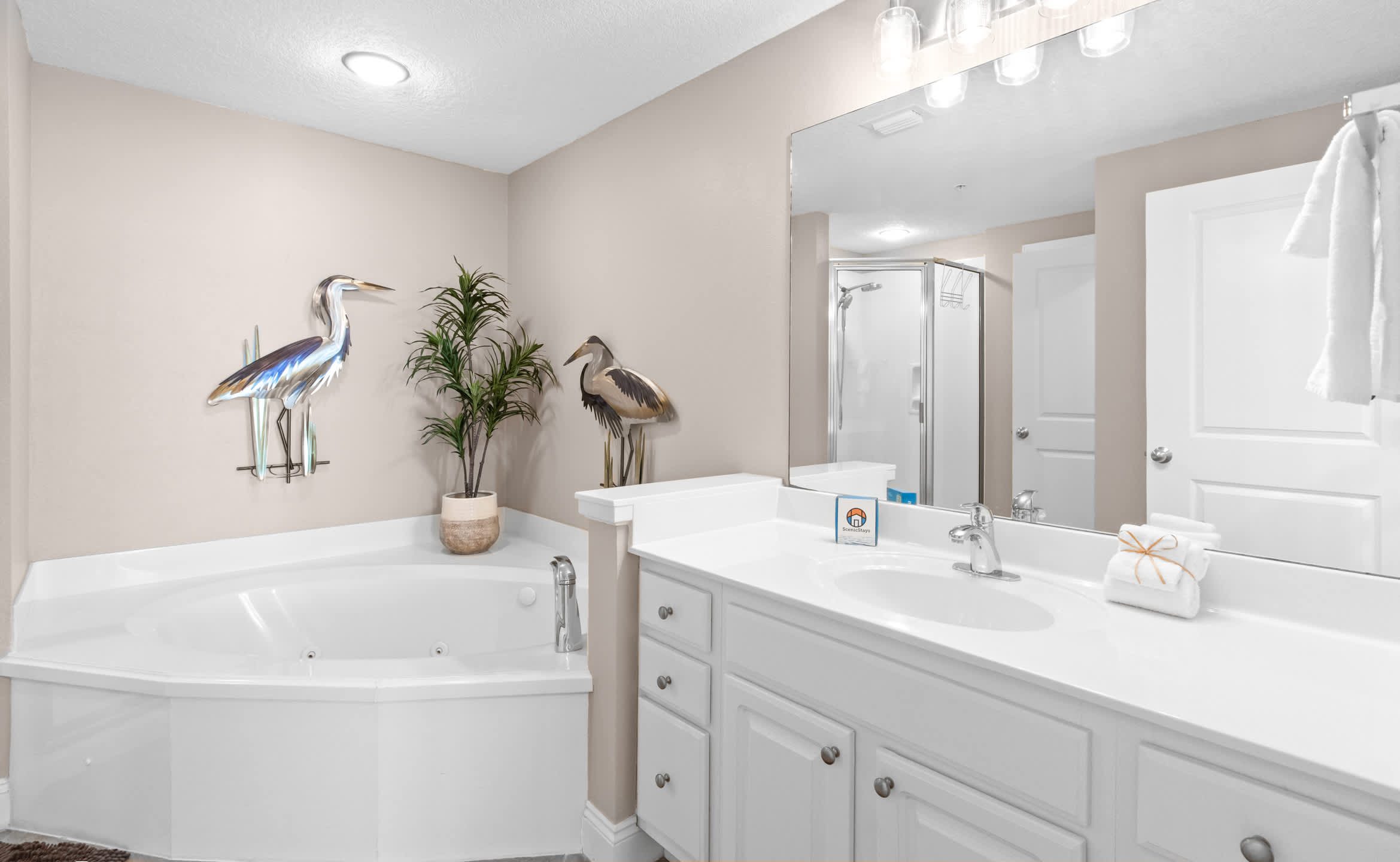 A spacious bathroom featuring a white single vanity, a corner jetted tub, and a separate glass-enclosed shower, all set against light beige walls and tan tiled flooring.