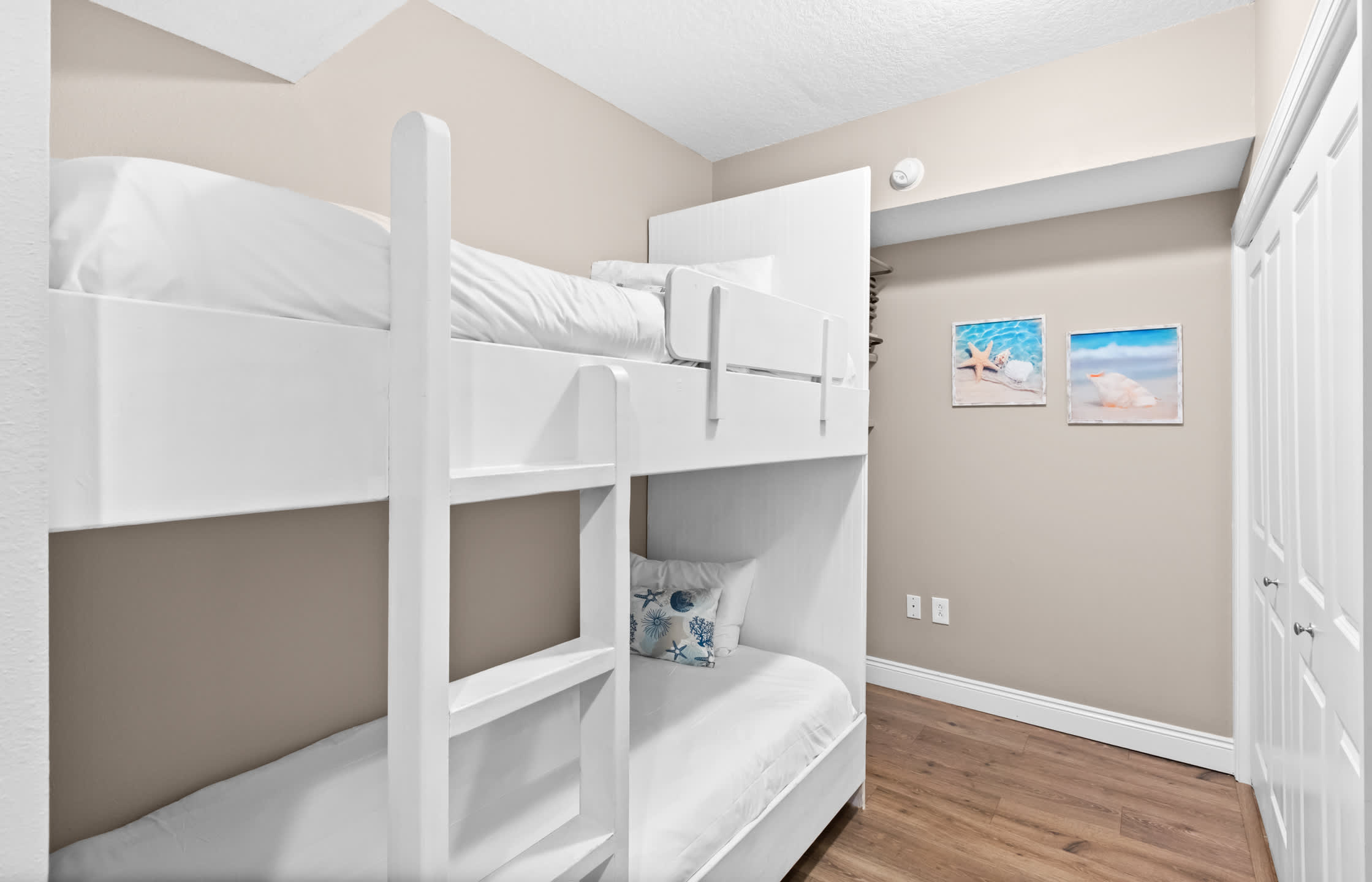 A cozy bedroom with a white wooden bunk bed, featuring white bedding and a decorative blue and white starfish pillow on the bottom bunk. The room has light beige walls, wood-look flooring, and two framed beach-themed pictures.