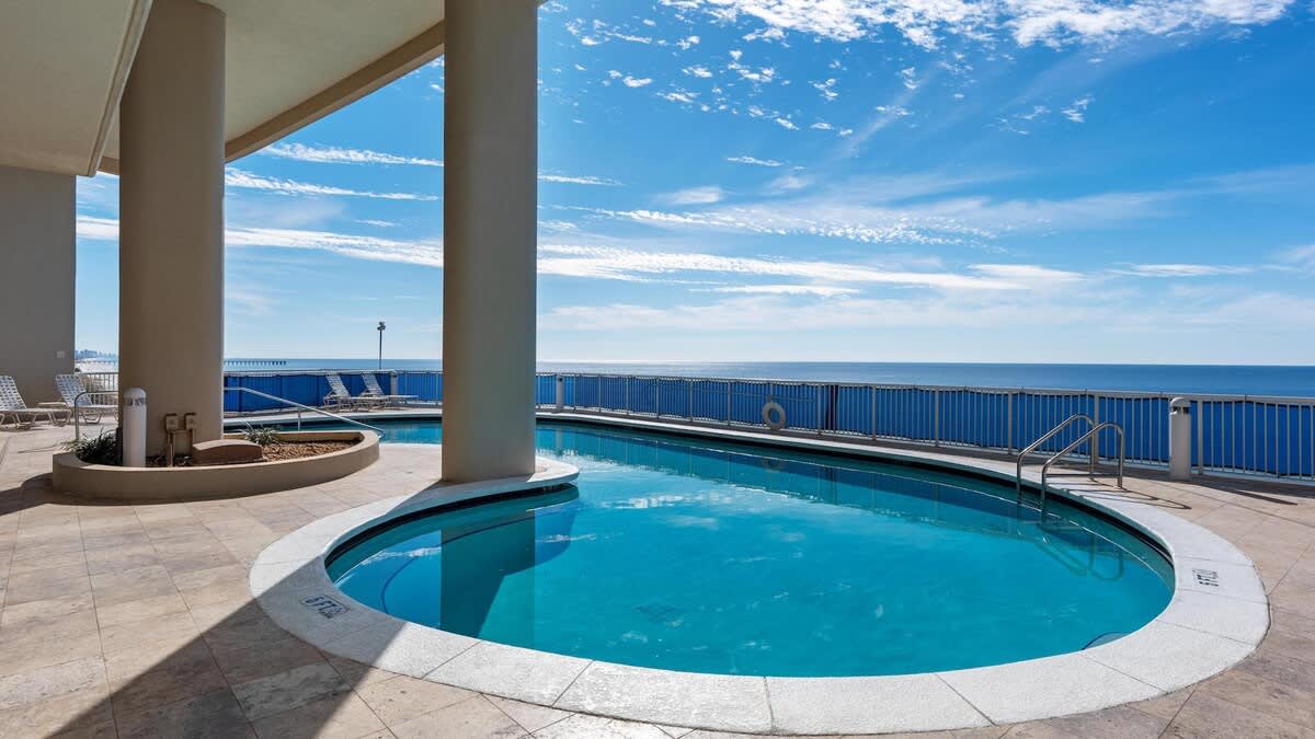 A kidney-shaped communal swimming pool with a light blue interior is set on a tiled deck, offering direct views of the expansive blue ocean under a bright, partly cloudy sky. Lounge chairs are visible in the background for relaxation.