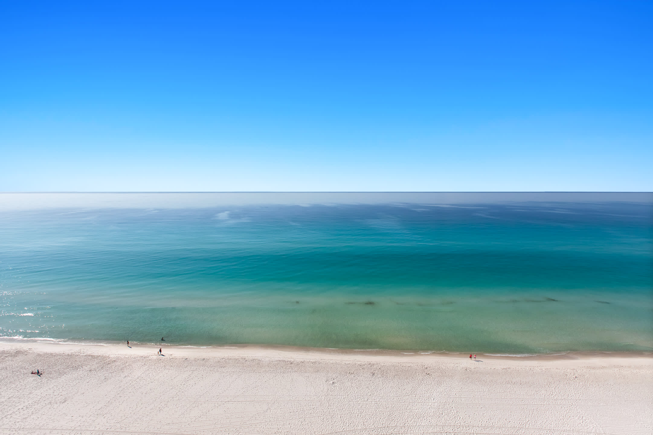A stunning aerial perspective showcases the vast expanse of a white sandy beach gently merging with the vibrant turquoise and deep blue hues of the calm ocean under a clear, bright sky. A few people are visible walking along the water's edge.