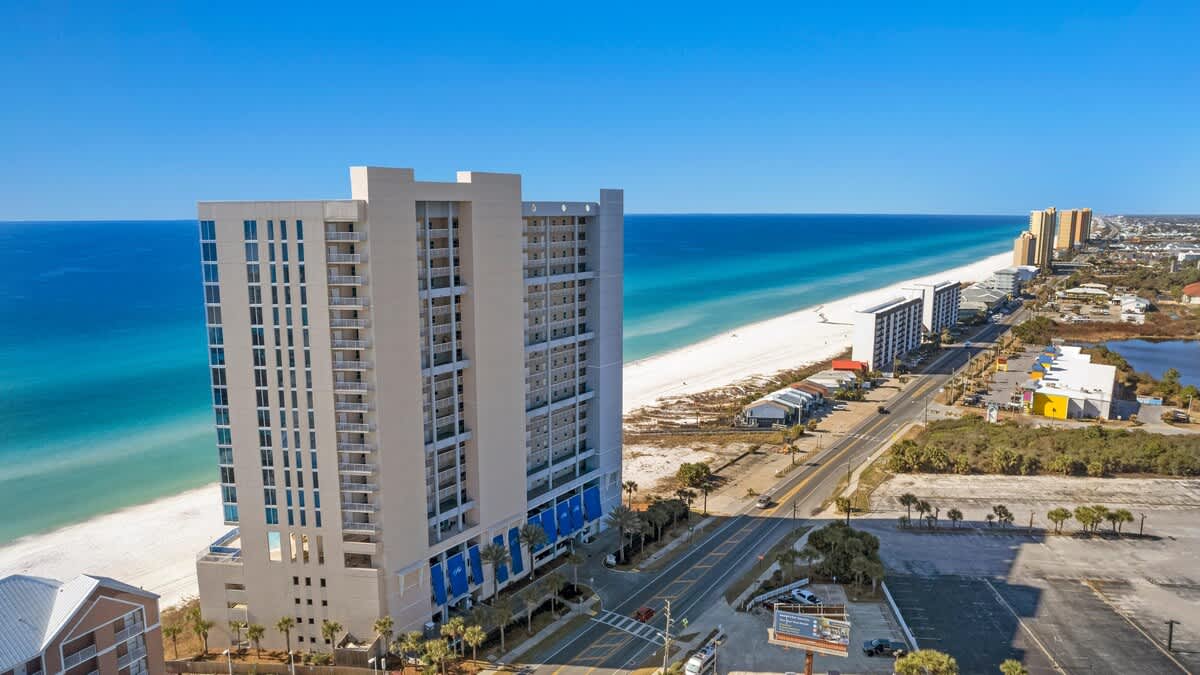 An aerial perspective highlights the tall, multi-story condominium building from a side angle, showcasing its numerous balconies overlooking a long stretch of white sand beach and the vibrant blue ocean. A coastal road runs parallel to the beach, with other buildings and coastal vegetation visible.