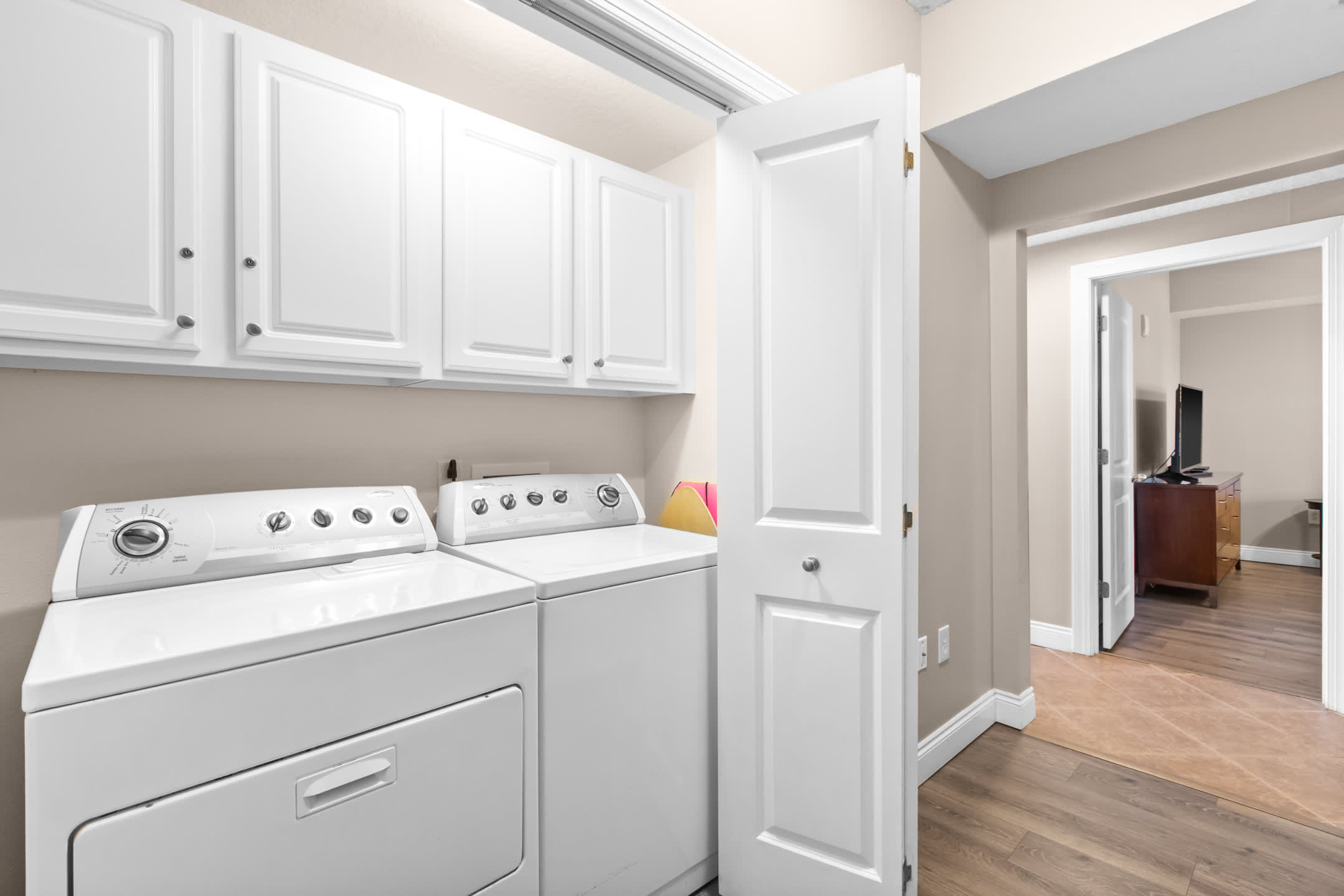 A dedicated laundry area featuring white top-loading washer and dryer units beneath white wall-mounted cabinets, set against light beige walls with wood-look flooring.
