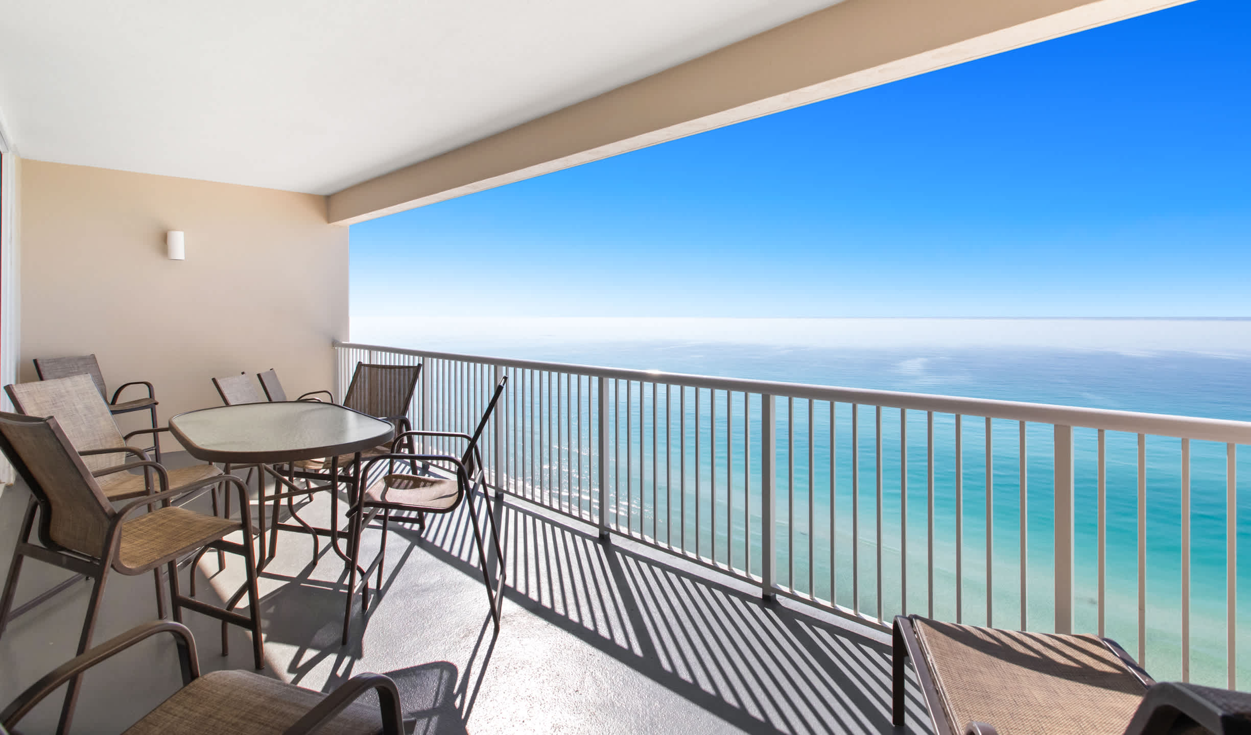 A spacious outdoor balcony with a panoramic view of the calm blue ocean and distant horizon, furnished with a glass-top table and several high-back chairs. The light gray floor and white railing frame the expansive seascape.
