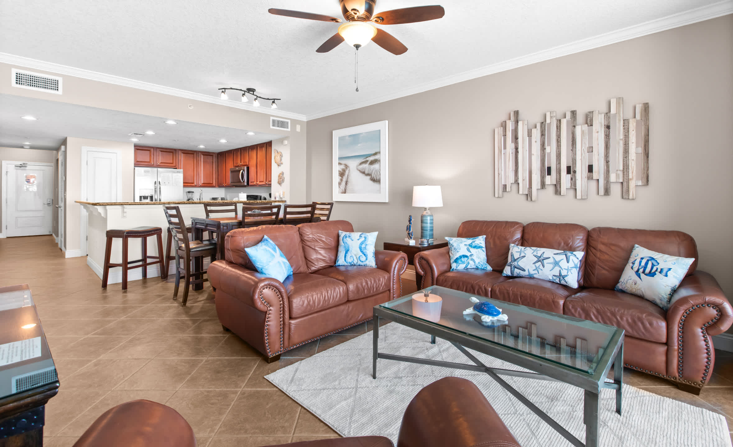 This bright open-concept space combines a living room with brown leather sofas and a glass coffee table, and a dining area with a wooden table, all set against light walls and tile flooring, with a clear view of the ocean through the balcony doors.