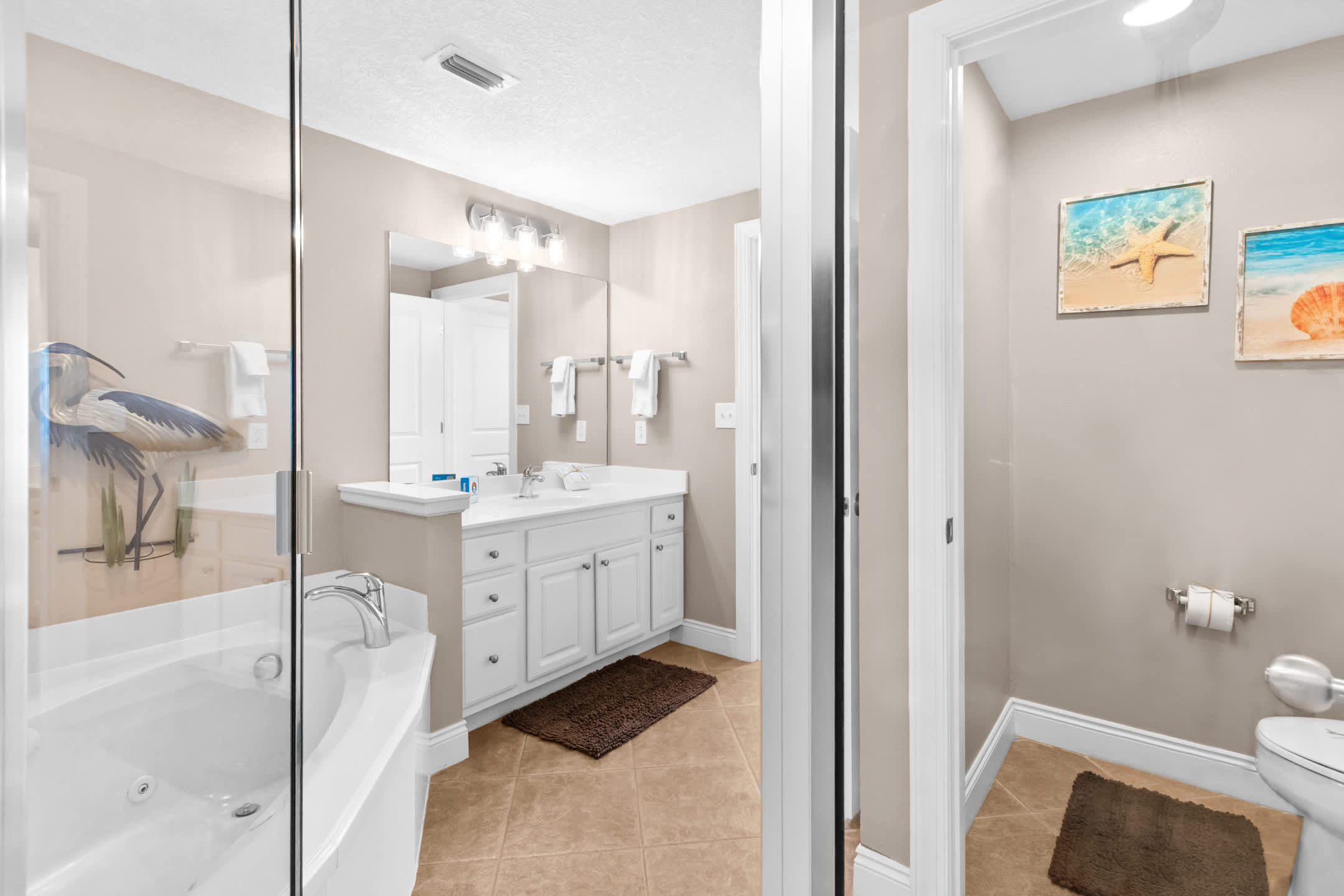 A bright bathroom with a double vanity featuring white cabinets and a light countertop, a large mirror, and a toilet visible in an adjacent alcove. The floor is tiled in a light brown, and the walls are light beige.