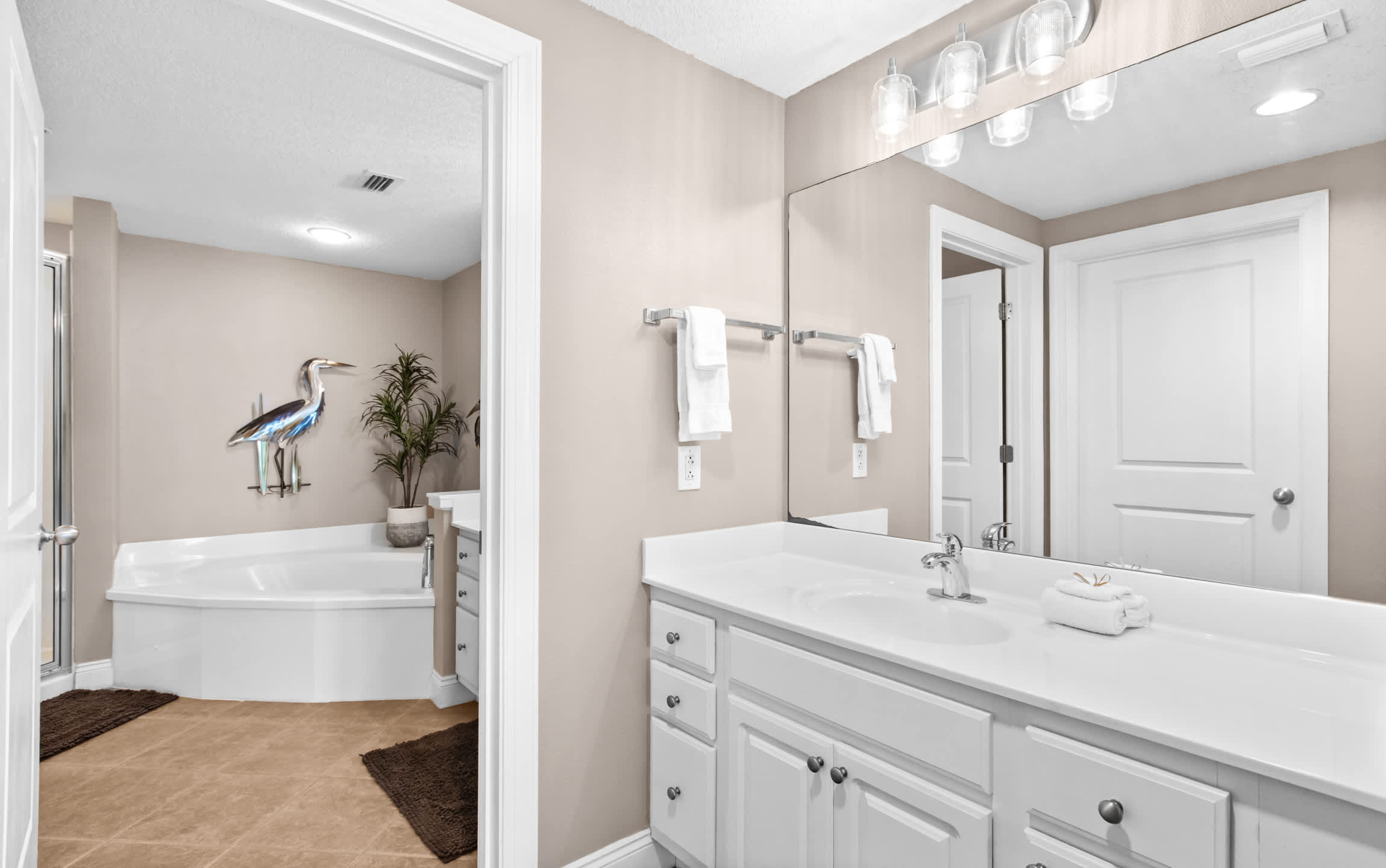 A well-lit bathroom with a white single vanity, a large mirror, and a corner jetted tub, accented by two decorative heron statues and a small plant.