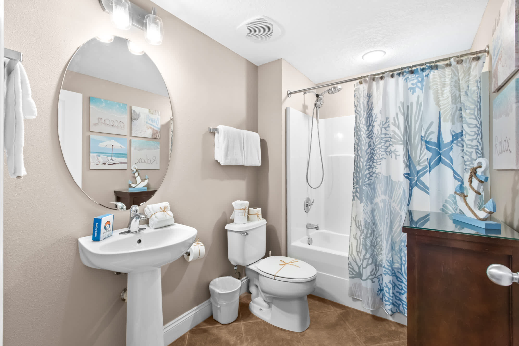 A bright bathroom featuring a white pedestal sink with a round mirror above it, a toilet, and a shower/tub combo with a blue and white coral-patterned shower curtain. The room has light beige walls and light brown tiled flooring.