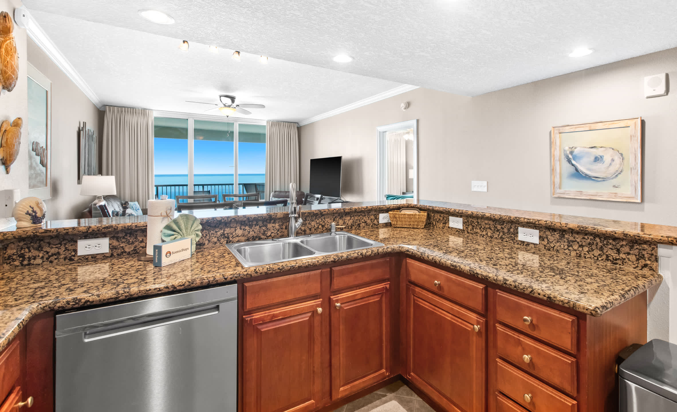 The kitchen's granite countertop extends to a double sink area, offering a view into the open-concept living room and balcony beyond, with stainless steel appliances and warm wooden cabinets completing the space.