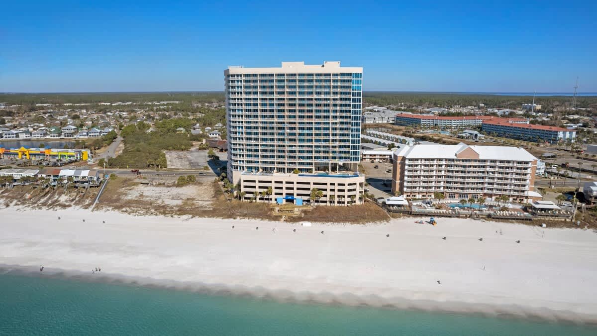 A wide aerial shot captures the full facade of a large, modern condominium building with numerous balconies, positioned directly on a pristine white sand beach. The clear blue ocean stretches into the distance, with other buildings and coastal development visible further inland.