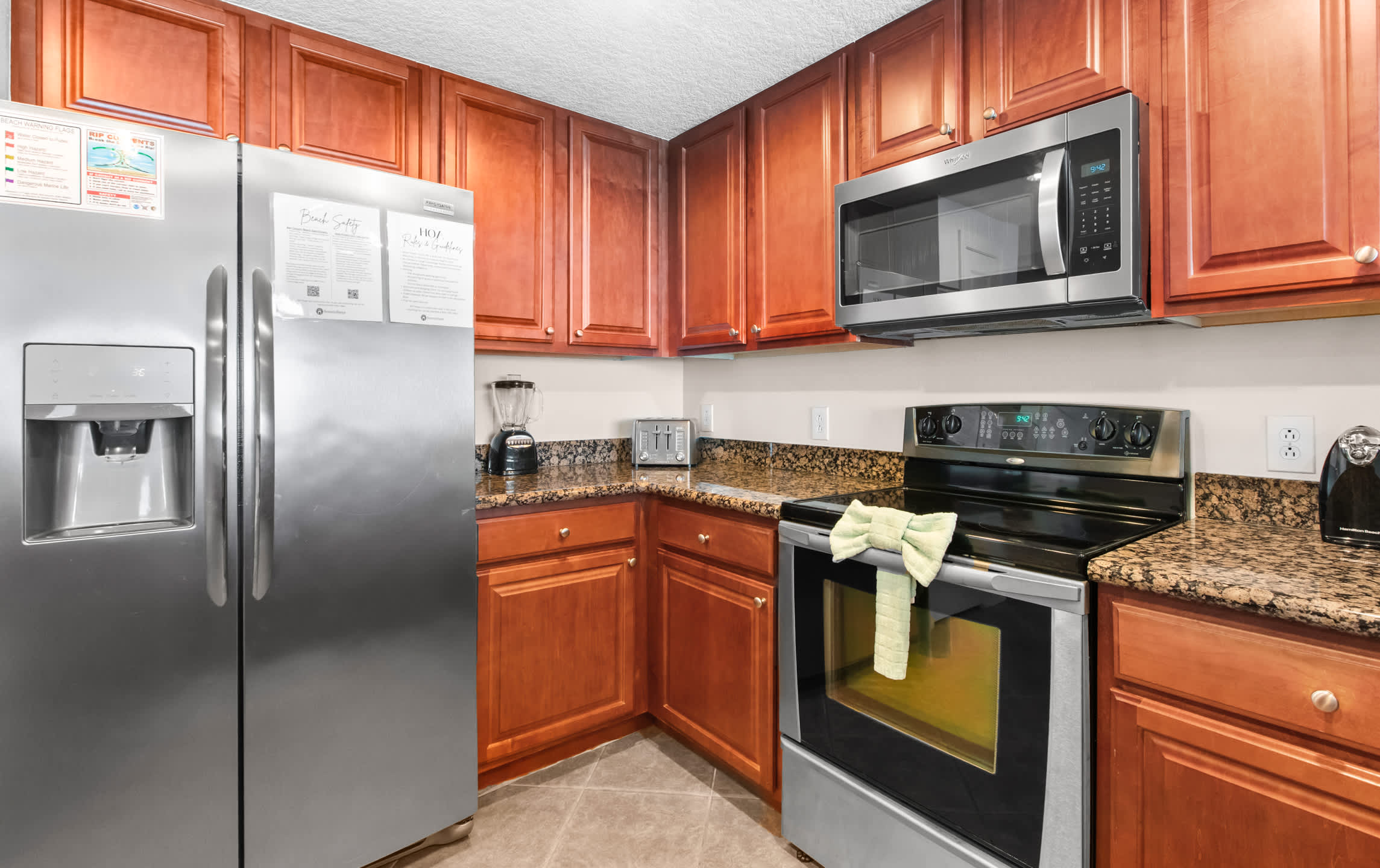 This kitchen corner features a large stainless steel French door refrigerator, ample granite counter space with a toaster and blender, and warm wooden upper and lower cabinets, providing a practical and inviting cooking area.