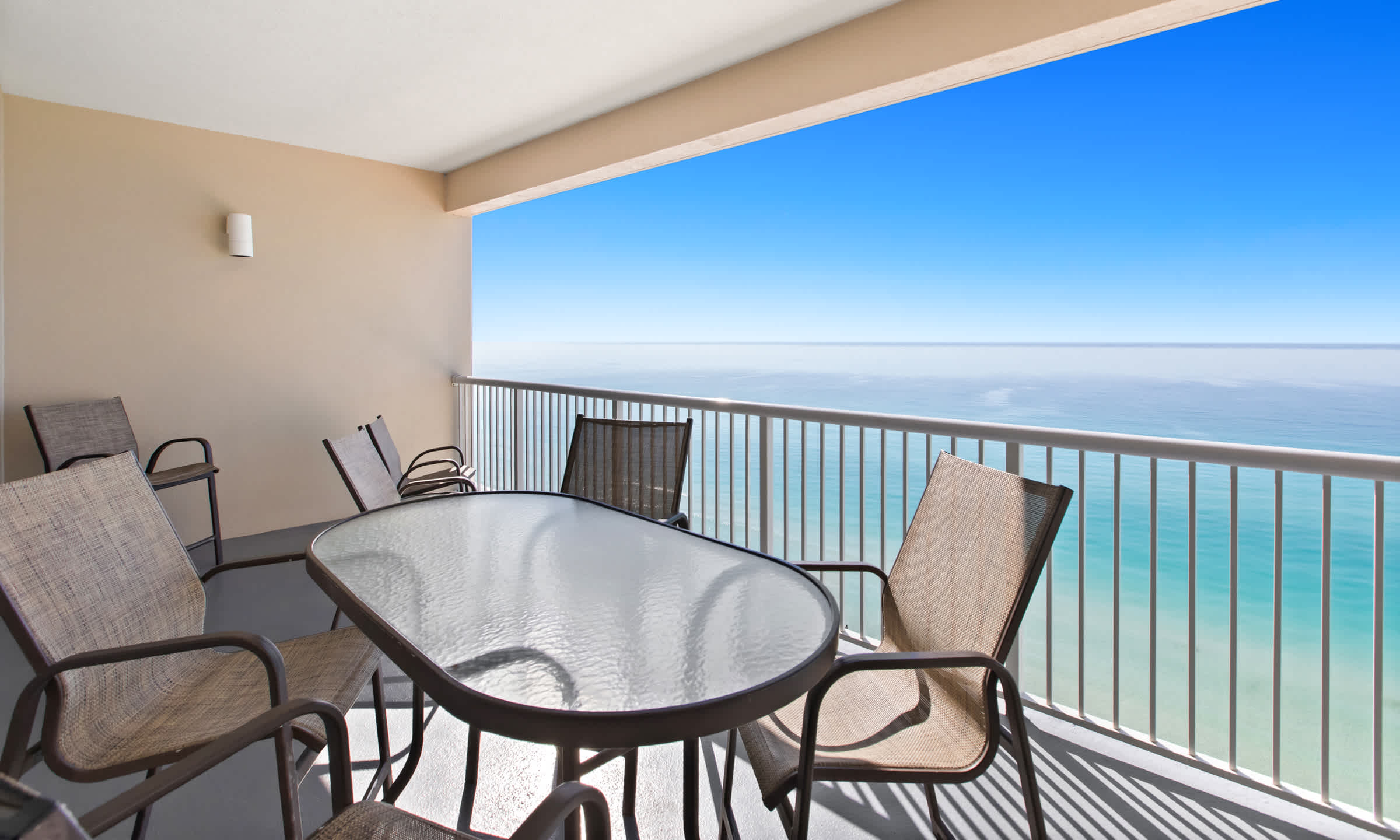 A wide outdoor balcony offering breathtaking views of the endless blue ocean and sky, equipped with a glass-top table and several chairs for outdoor dining. Sunlight casts long shadows across the light gray floor.