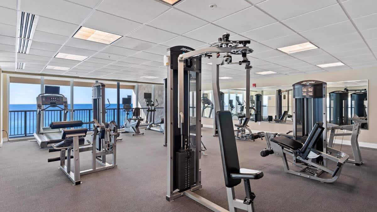 A well-equipped fitness center features a variety of modern exercise machines, including treadmills, ellipticals, and weight stations, set on dark carpeted flooring. Large windows offer panoramic ocean views, creating an inspiring workout environment.