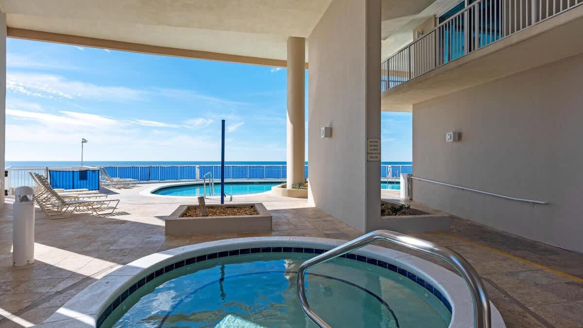 An inviting outdoor hot tub with blue tile accents is situated under a covered patio, adjacent to a communal pool. Beyond the railing, the vast blue ocean stretches to the horizon under a bright sky, offering a relaxing waterside experience.