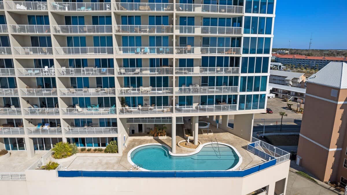 An aerial perspective looks down on the property's outdoor pool deck, featuring a uniquely shaped swimming pool, situated on a lower level of the tall, modern condominium building. Multiple balconies with blue-tinted windows overlook the pool area and the surrounding landscape.