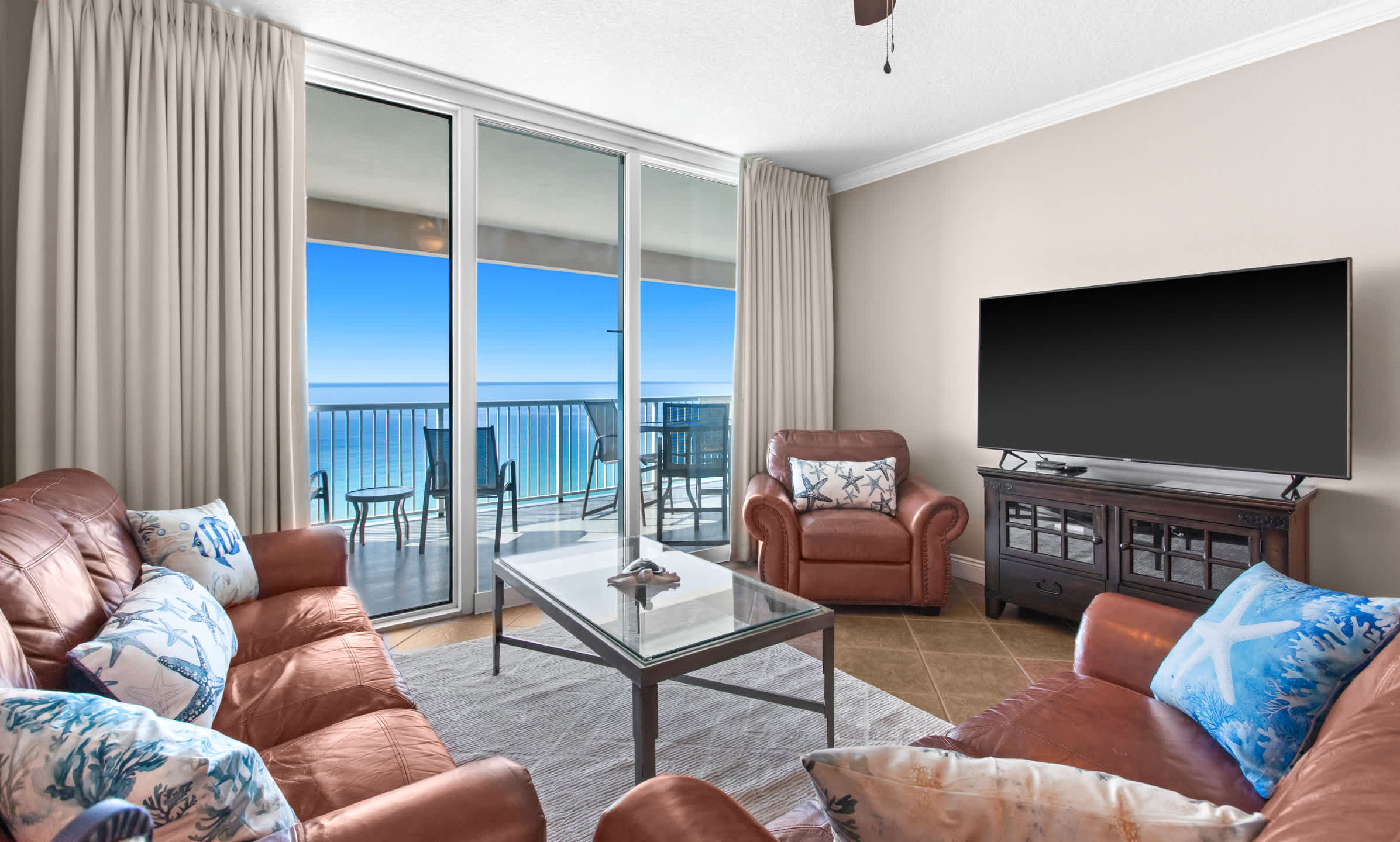 This inviting living room showcases brown leather seating, including two sofas and an armchair, arranged around a glass coffee table on a light rug, with a large flat-screen TV on a dark wood console and expansive ocean views through the balcony doors.