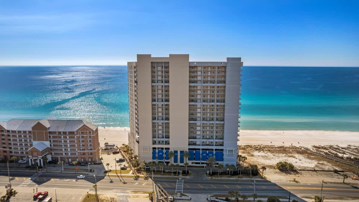 An aerial shot captures the multi-story condominium building from the front, featuring a modern design with many balconies, positioned directly on the white sandy beach. The vibrant blue ocean is visible behind it, with a road and other buildings in the foreground.
