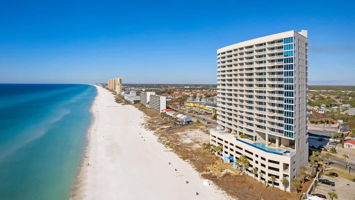 A striking aerial view showcases the tall, modern condominium building with numerous balconies, standing prominently alongside a wide, white sandy beach. The turquoise ocean stretches into the distance under a clear blue sky, with other buildings visible along the coastline.