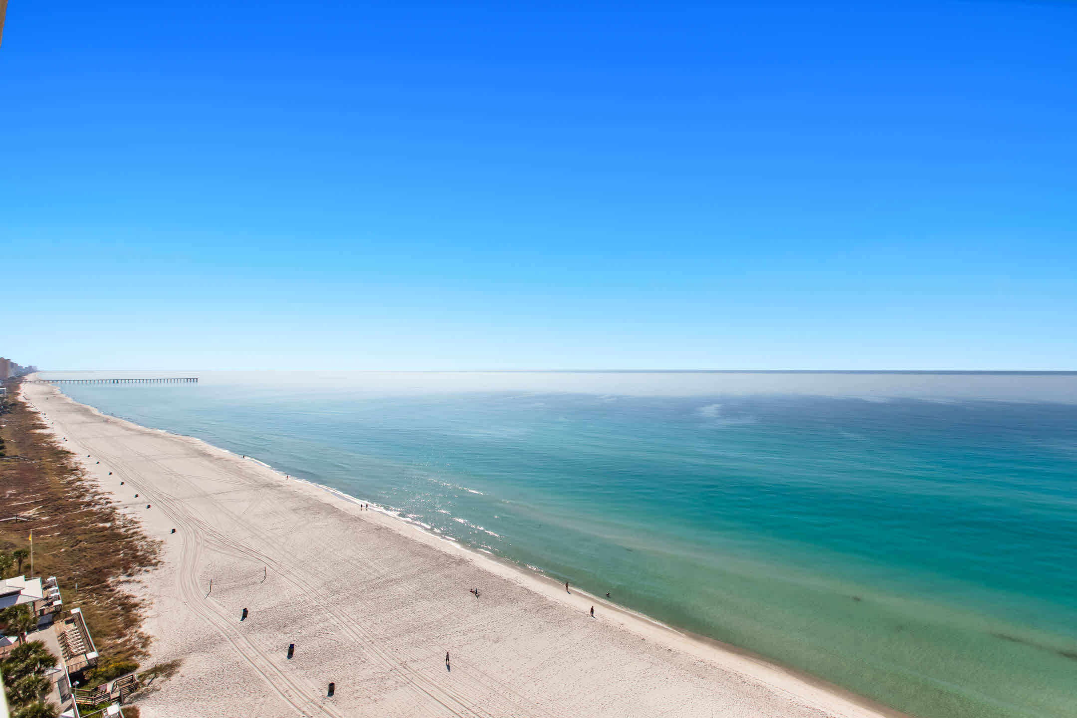 An expansive aerial view captures a wide, pristine sandy beach meeting the clear turquoise waters of the ocean under a bright blue sky, with a long pier extending into the hazy distance. Scattered individuals enjoy a leisurely stroll along the shoreline.