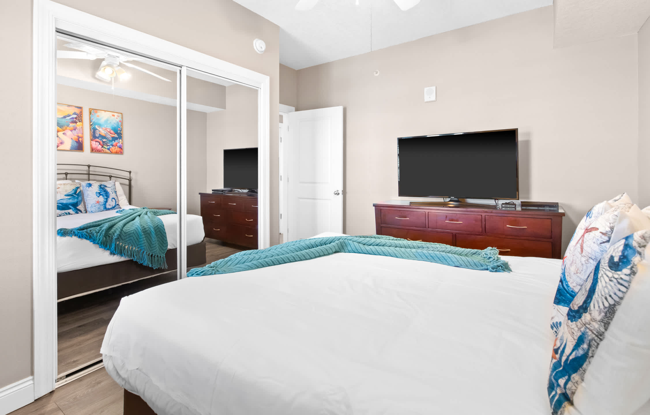 View of bedroom-1 showing a dark wood dresser with a flat-screen TV, mirrored closet doors reflecting the bed, and light wood-look flooring.
