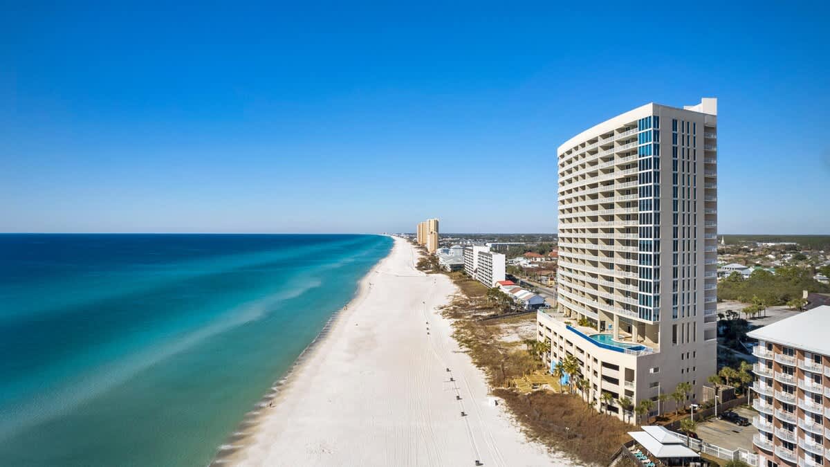 An expansive aerial shot reveals the long, pristine white sand beach and the brilliant blue ocean, with a tall, modern condominium building featuring multiple balconies positioned on the right. The view extends along the coastline, showing other distant buildings and the clear horizon.
