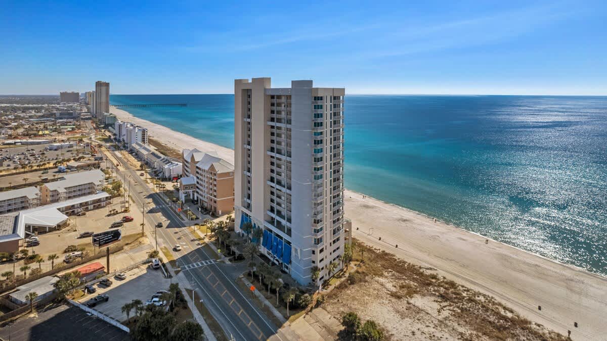 A wide aerial view captures the tall, contemporary condominium building situated between a bustling coastal road and a pristine white sandy beach. The expansive turquoise ocean stretches to the horizon under a bright blue sky, with a long pier visible in the distance.