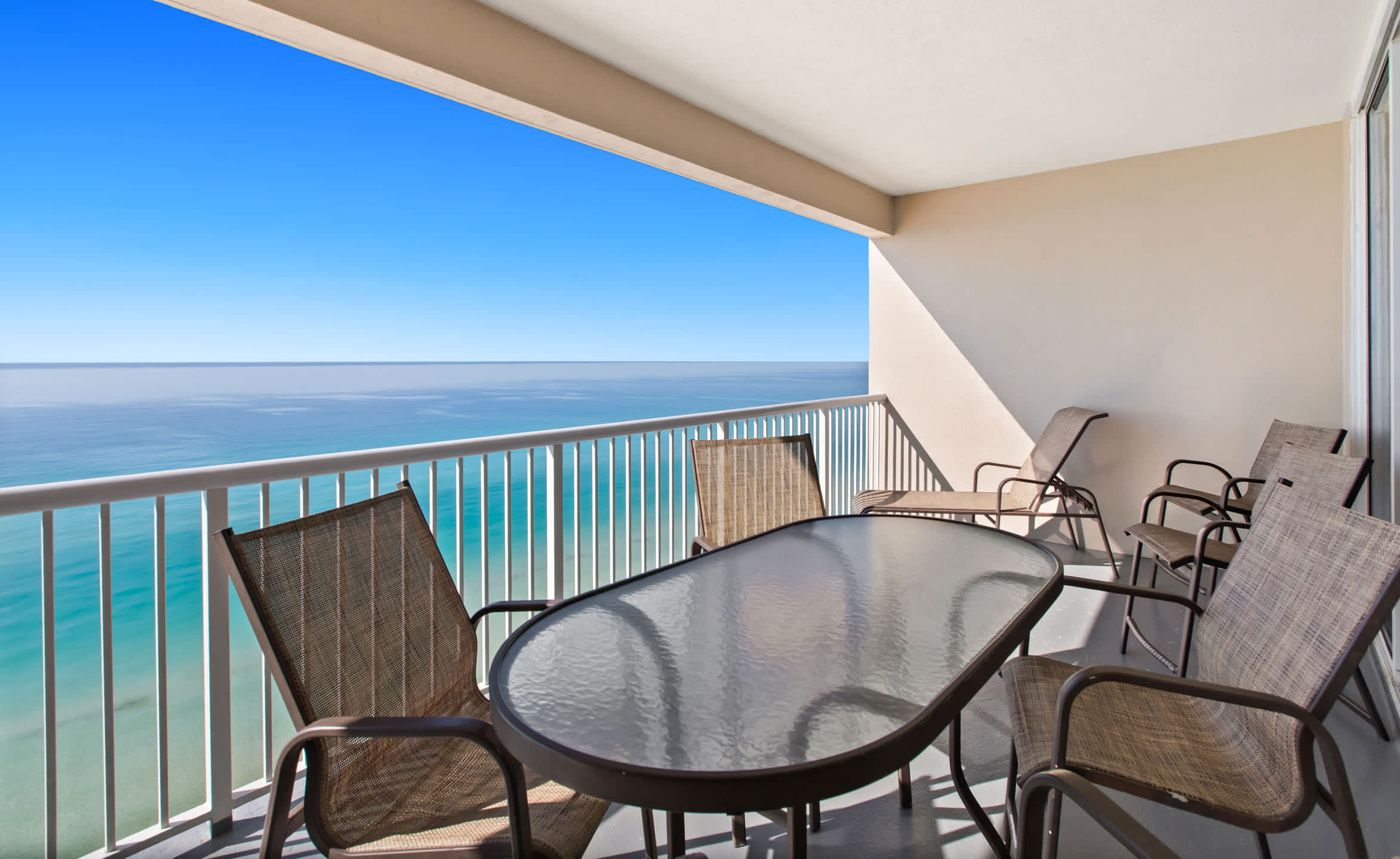 An expansive outdoor balcony with stunning views of the turquoise ocean and horizon, featuring a glass-top dining table and multiple mesh-backed chairs. The light gray floor and white railing complement the bright blue sky.