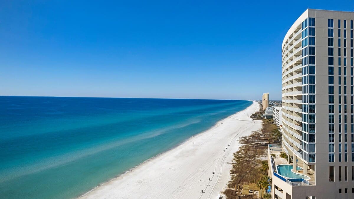 A breathtaking aerial view captures the vast expanse of a white sand beach meeting the clear turquoise ocean, with a modern condominium building featuring a communal pool on its lower level situated on the right. The coastline stretches far into the horizon under a bright blue sky.
