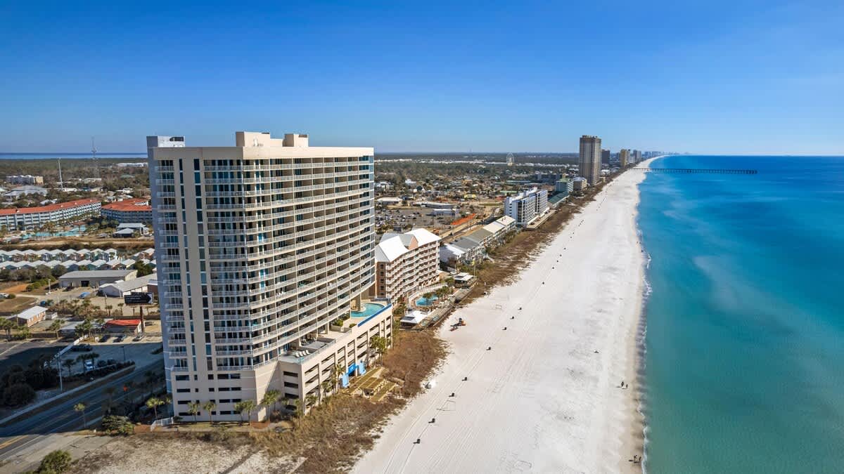 An aerial view showcases a tall, modern condominium building with multiple balconies, situated directly on a wide, white sand beach next to the turquoise ocean. A communal pool is visible on a lower level, with other resort buildings and a town in the background.
