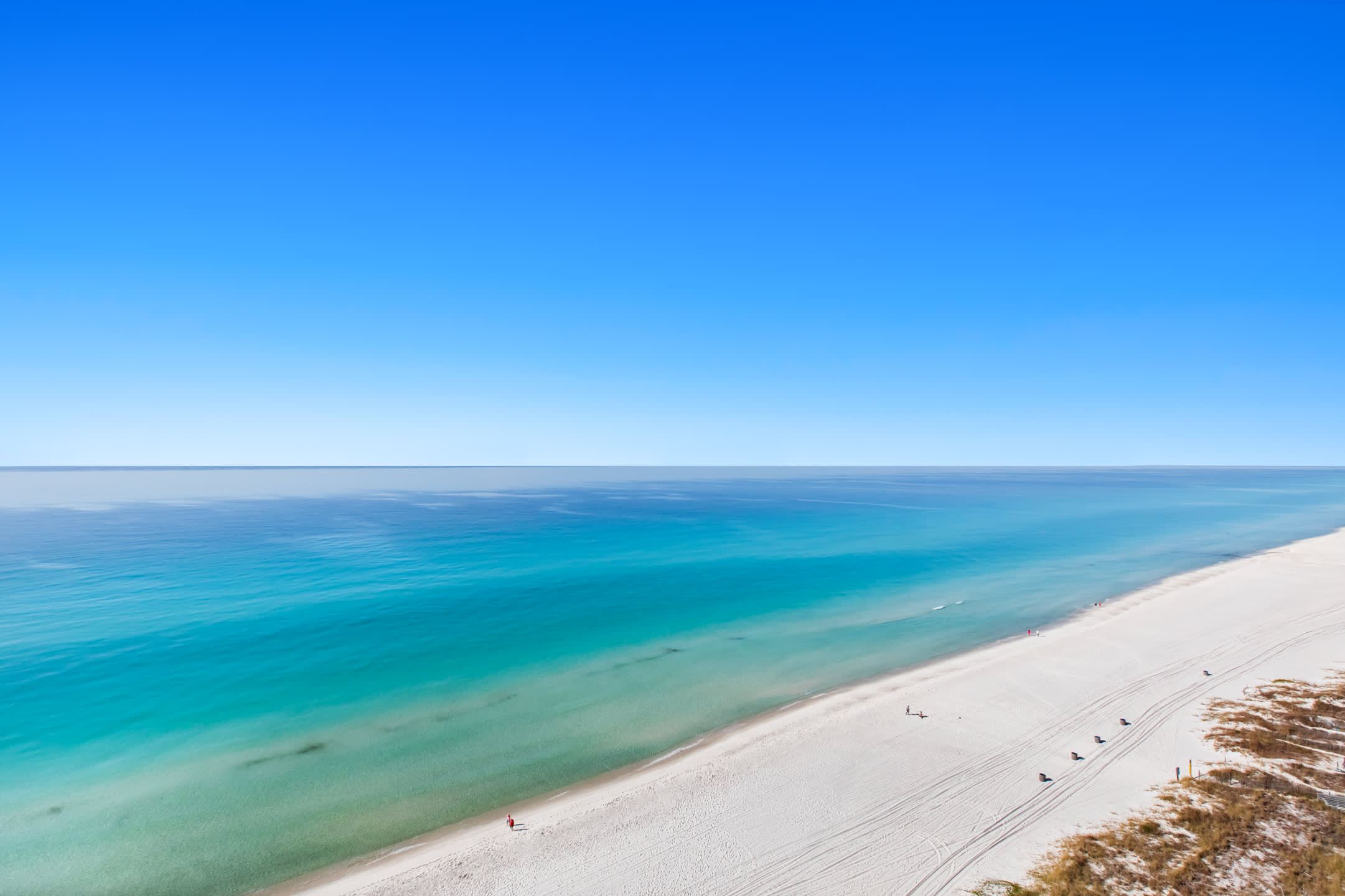 An elevated view reveals a long stretch of white sandy beach bordered by the clear, shallow turquoise waters of the ocean, transitioning to deeper blue. Sparse coastal vegetation and dunes line the right side, with a few people enjoying the sunny shoreline.