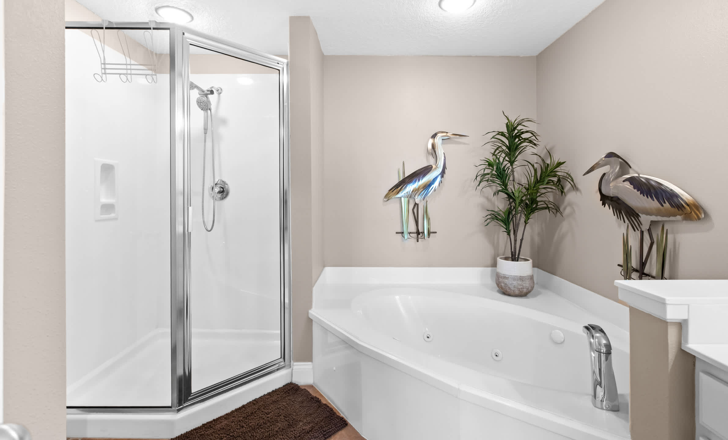 A spacious bathroom featuring a glass-enclosed shower and a corner jetted bathtub, accented with two large heron sculptures and a potted plant. The walls are light beige, and the floor has a brown bath mat.