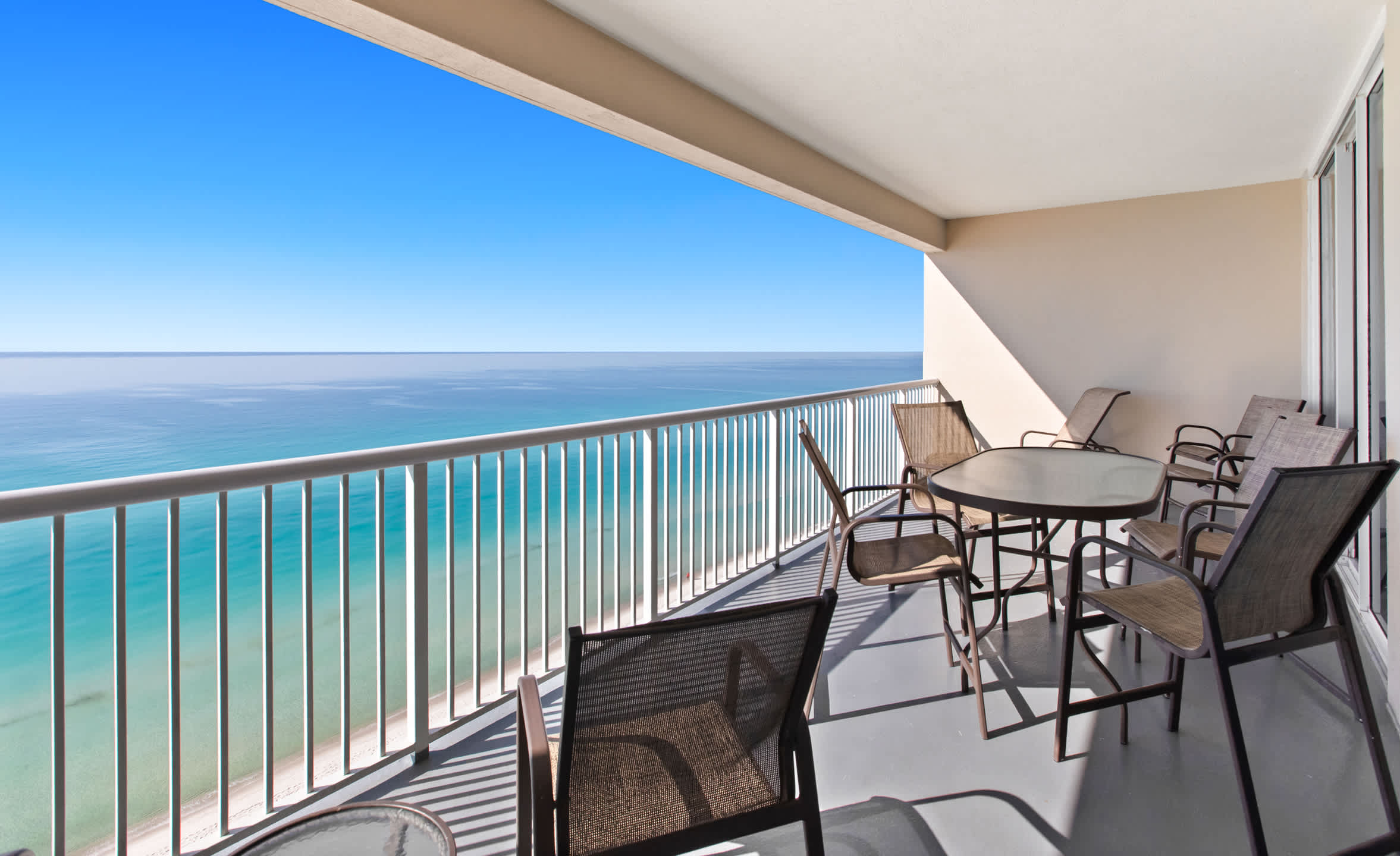 A spacious outdoor balcony overlooking a vast blue ocean, furnished with a glass-top dining table and several chairs. The railing is white, and the floor is a light gray, offering panoramic views of the coastline.