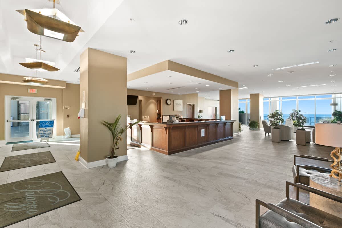 Spacious and well-lit lobby area featuring a large wooden reception desk and light-colored tiled floors. Seating areas and expansive windows offering panoramic ocean views are visible in the background.