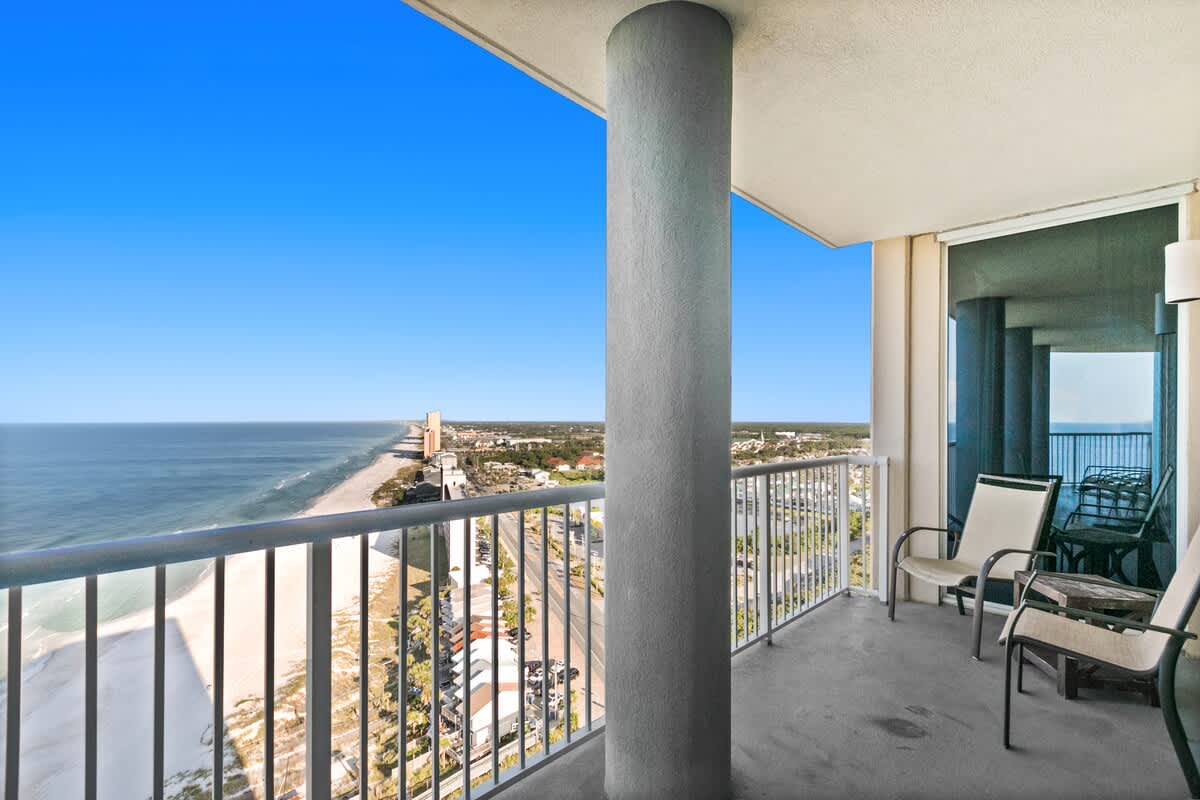 Expansive concrete balcony with a metal railing, offering panoramic views of the ocean, beach, and coastal city skyline under a clear blue sky. A large concrete pillar stands prominently on the left.