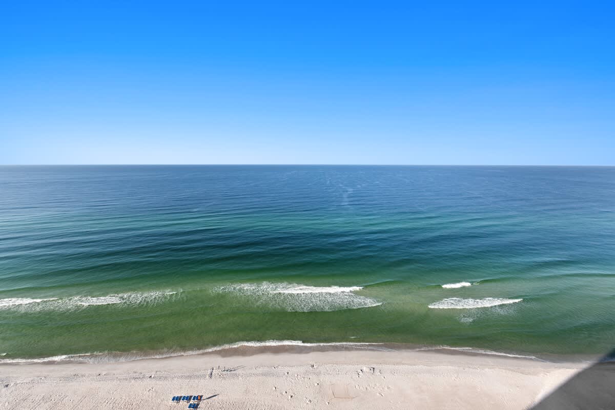 Straight-on, high-angle view of the wide sandy beach and the clear blue-green ocean with white waves breaking near the shore. The horizon is perfectly flat under a bright blue sky.