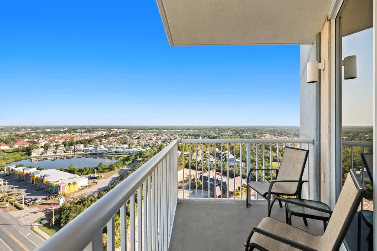 Outdoor balcony furnished with two chairs and a small table, offering a panoramic high-rise view of a city, a serene lake, and distant buildings under a clear blue sky.