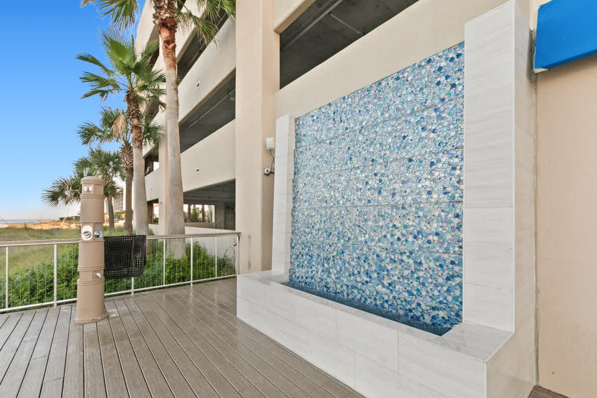 Outdoor deck area featuring a modern water wall with blue mosaic tiles, set against a backdrop of lush palm trees. A wooden walkway leads towards the beach access, offering a refreshing ambiance.
